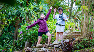 Two smiling women wave while standing on a rock path in a lush, green garden in Costa Rica.