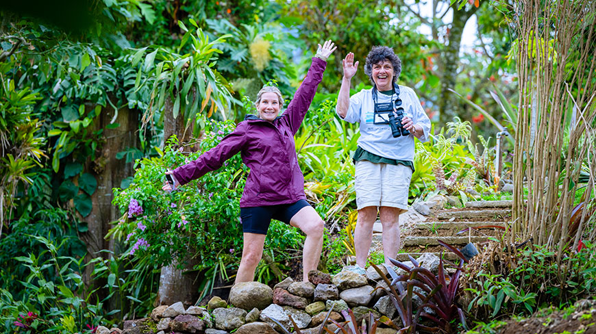 Two women wave happily while standing on a stone path in a lush garden in Costa Rica.