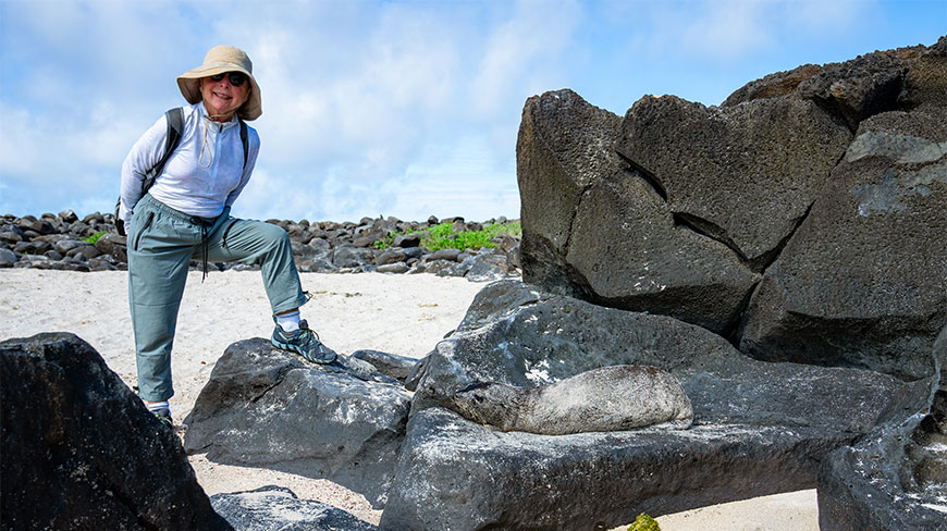 A woman in hiking gear stands near a sea lion resting on volcanic rocks during a hike at Punta Suarez, Espanola, Galapagos.