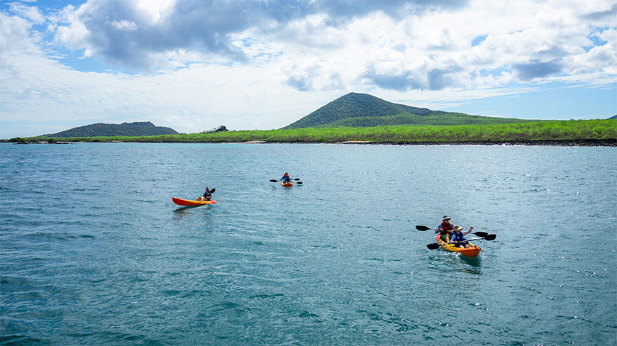 People kayaking in the blue waters of Baroness Point, Floreana Island, Galapagos, with lush green hills in the background.