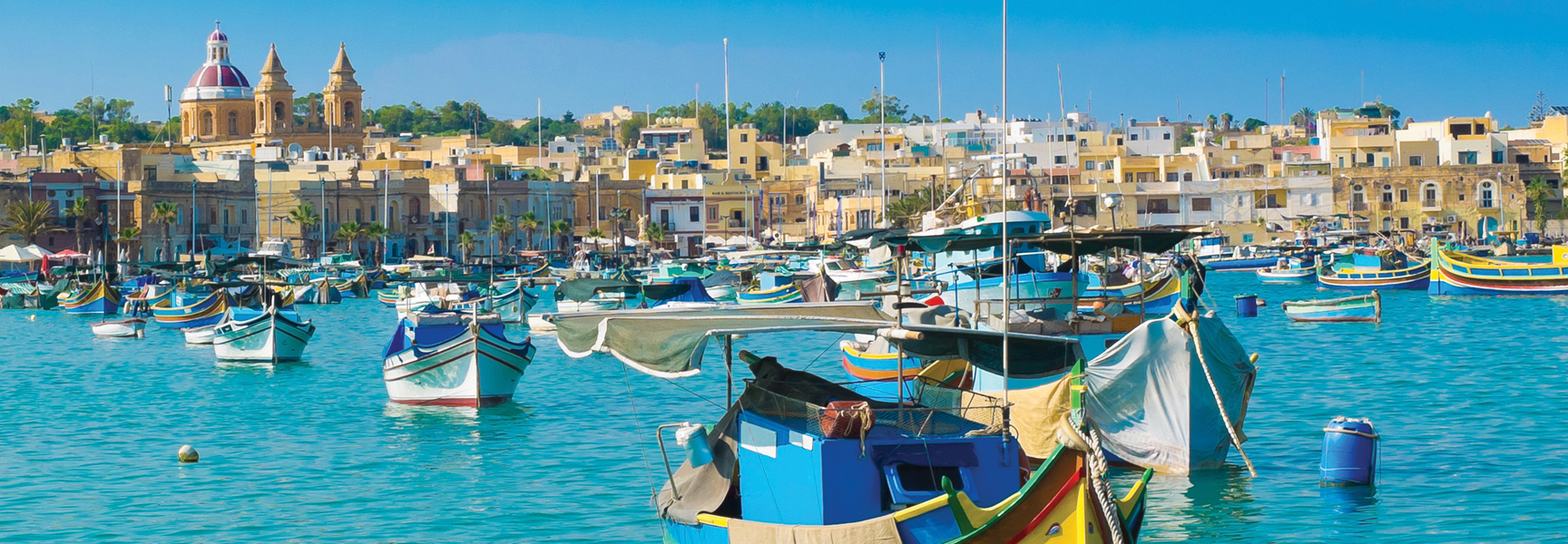 Colorful fishing boats float in the bright blue harbor of a seaside town in Malta under a clear sky.