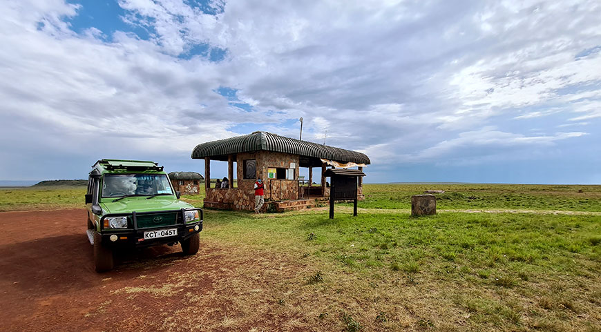 A green safari jeep is parked by a stone ranger station at the entrance to a vast, grassy plain.
