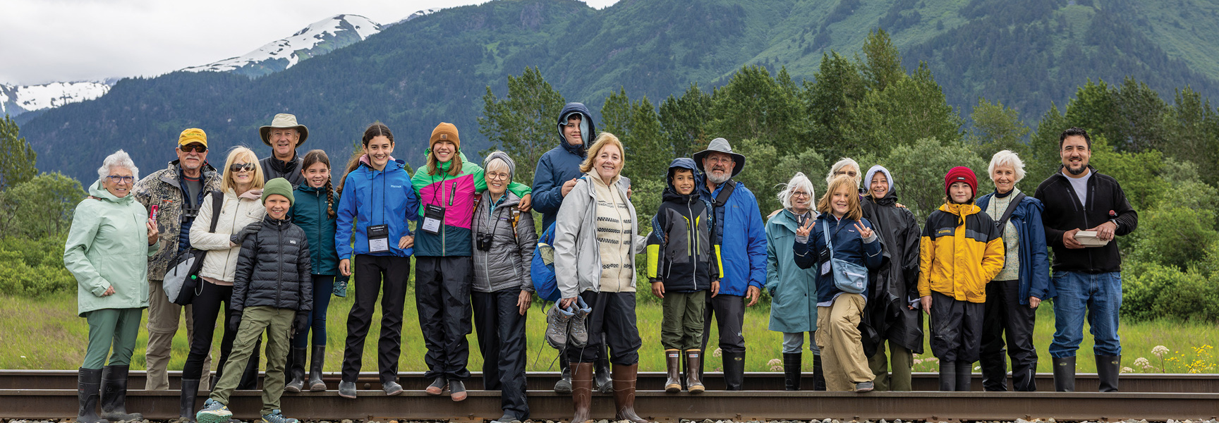 A large group of travelers of all ages pose for a photo on railroad tracks in front of snowy mountains in Alaska.