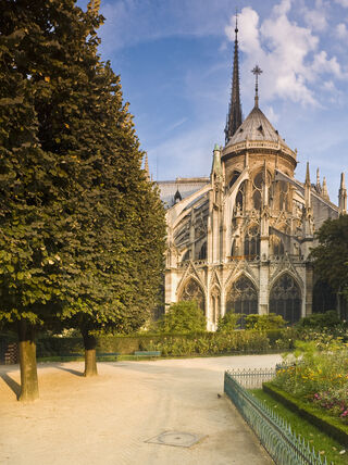 A panoramic view of the Notre-Dame Cathedral in Paris from a lush garden with colorful flowers and a gothic fountain under a clear sky.