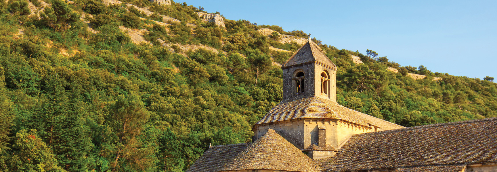 A historic stone cathedral with a bell tower is nestled into a sun-drenched, tree-covered hillside in Provence.