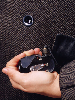 A hand holds a concealed spy camera with a cable leading to a hidden lens disguised as a button on a herringbone tweed coat.