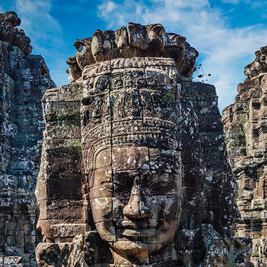 Three ancient, weathered stone towers at Angkor Wat in Cambodia feature large, serene faces carved into the rock under a blue sky.