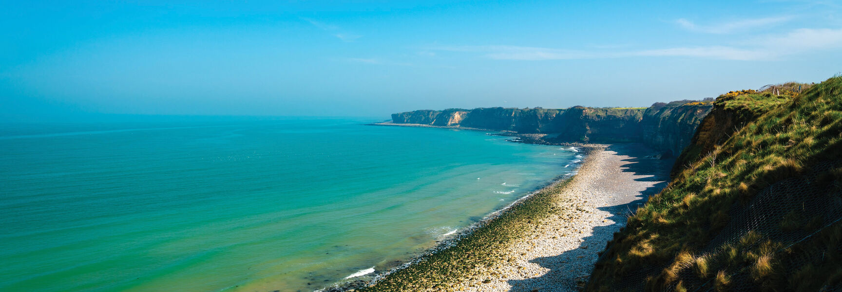 Wide view of the Normandy Beaches showing steep coastal cliffs and a rocky shoreline bordering turquoise waters under a bright blue sky.
