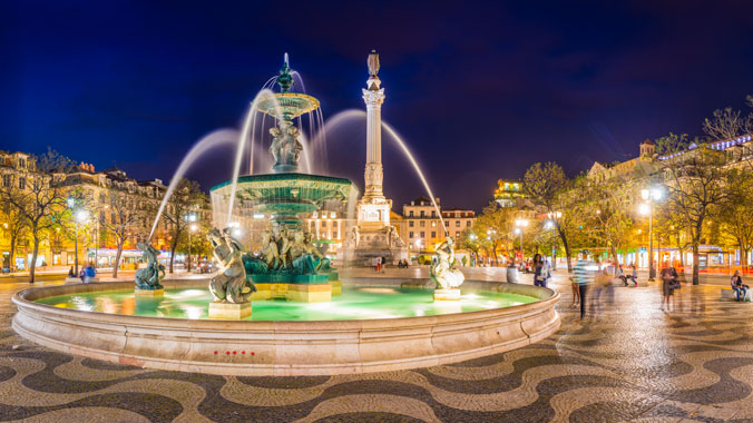 The ornate fountain in Lisbon's Rossio Square is illuminated at night, with the Column of Pedro IV and distinctive wave-patterned cobblestones visible.