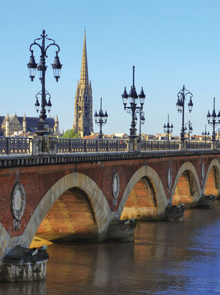 An arched stone bridge with ornate black lampposts stretches across a river in Bordeaux, France, with a tall cathedral spire in the background.