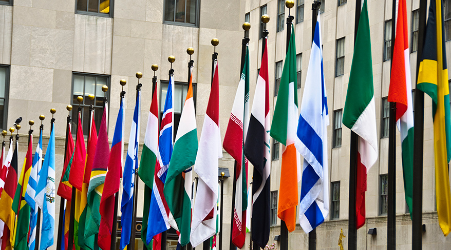 A row of colorful international flags flies on flagpoles in front of a stone building.