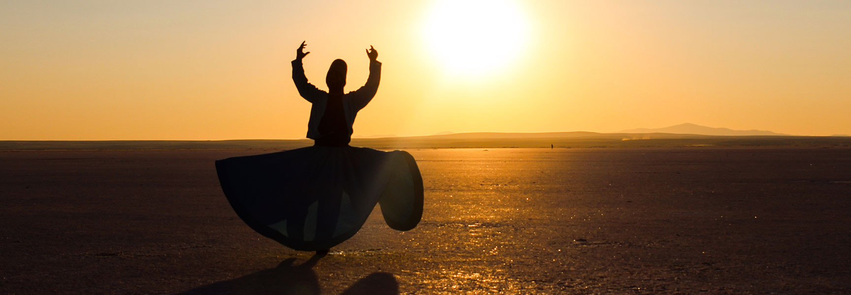 A silhouetted figure of a whirling dervish spins in a large, open salt flat at sunset.