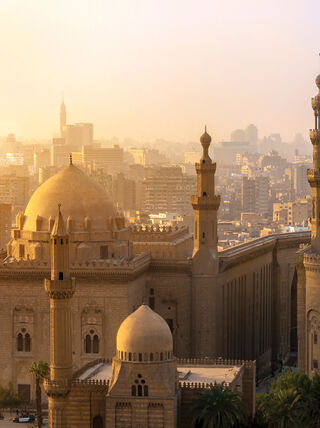 A golden sunset glows over the historic mosques and minarets of Cairo, Egypt, highlighting the intricate architecture against a hazy city skyline.