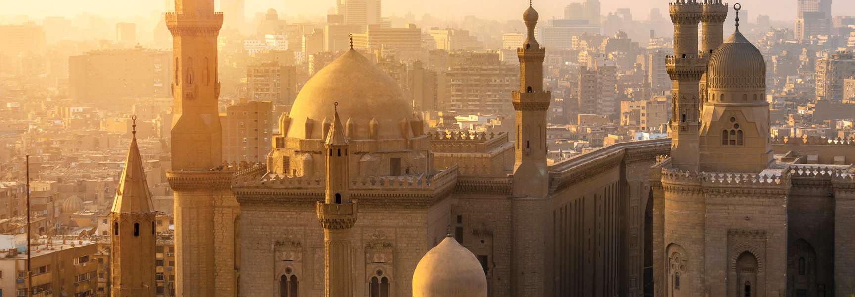 The domes and minarets of historic mosques in Cairo, Egypt, are bathed in the golden light of sunset with the city skyline behind.