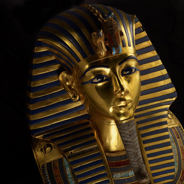 The ornate gold funerary mask of King Tutankhamun featuring blue lapis lazuli stripes and a false beard against a dark background.
