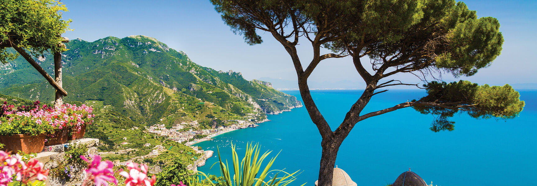 A vibrant landscape of the Amalfi Coast in Italy featuring a stone pine tree and pink flowers overlooking the blue Mediterranean Sea and mountains.