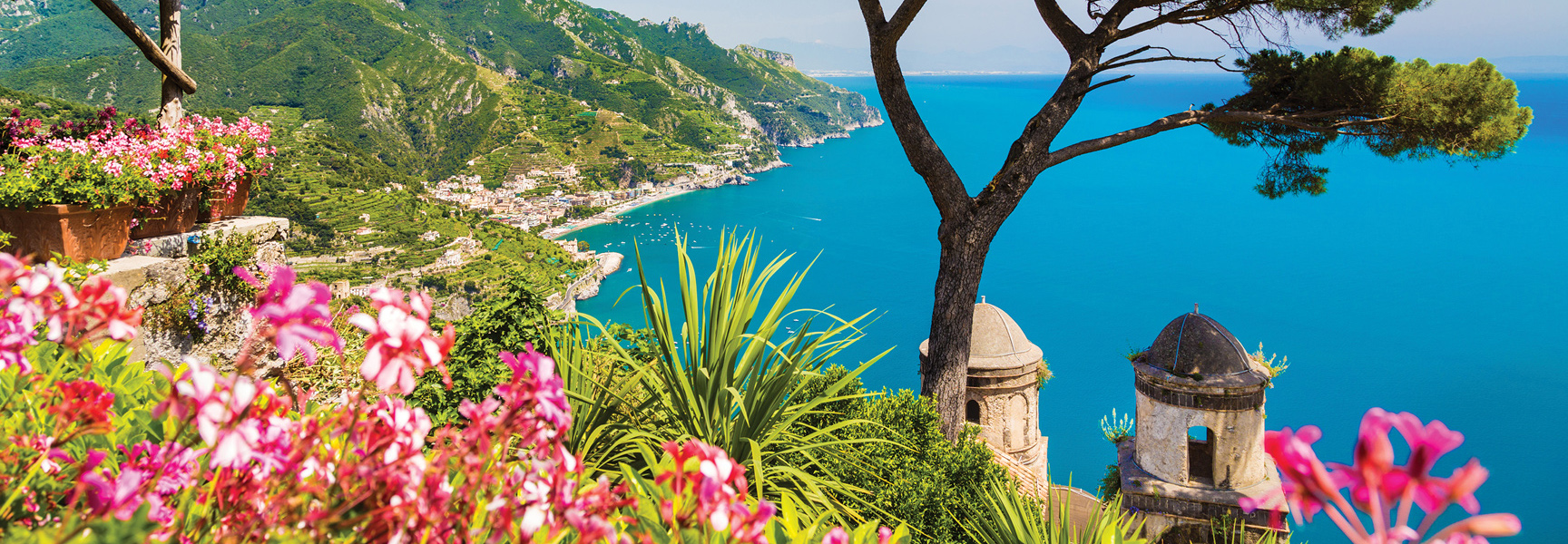A sunny, high-angle view of the Amalfi Coast in Italy, looking past pink flowers to the terraced green cliffs and brilliant blue sea.