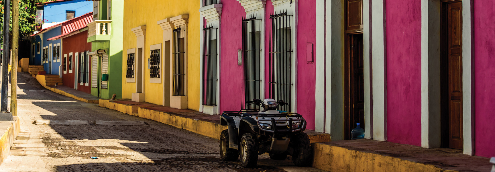 An ATV is parked on a quiet, sunlit street next to brightly colored buildings in a town in Mexico.