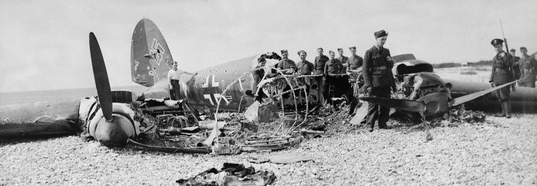 A black and white photo shows soldiers inspecting the wreckage of a downed German aircraft during the Battle of Britain.