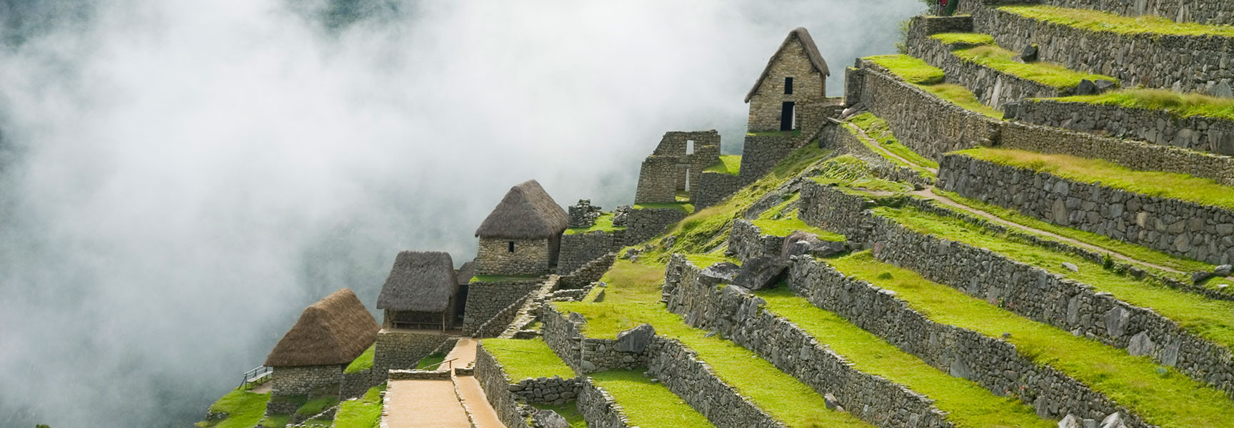 Ancient Incan stone buildings and green terraces of Machu Picchu emerge from a thick layer of clouds on a mountainside.