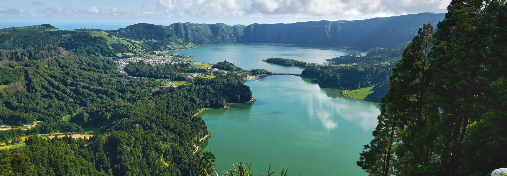 A panoramic view of the Sete Cidades crater lake in the Azores, surrounded by lush green volcanic hills, a bridge, and a small lakeside village.