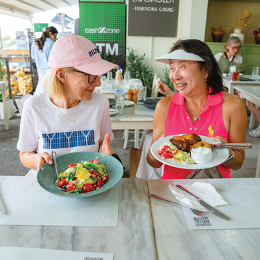 Two women smile while displaying plates of local cuisine at a restaurant in Greece or Turkey during a gastronomical learning adventure.