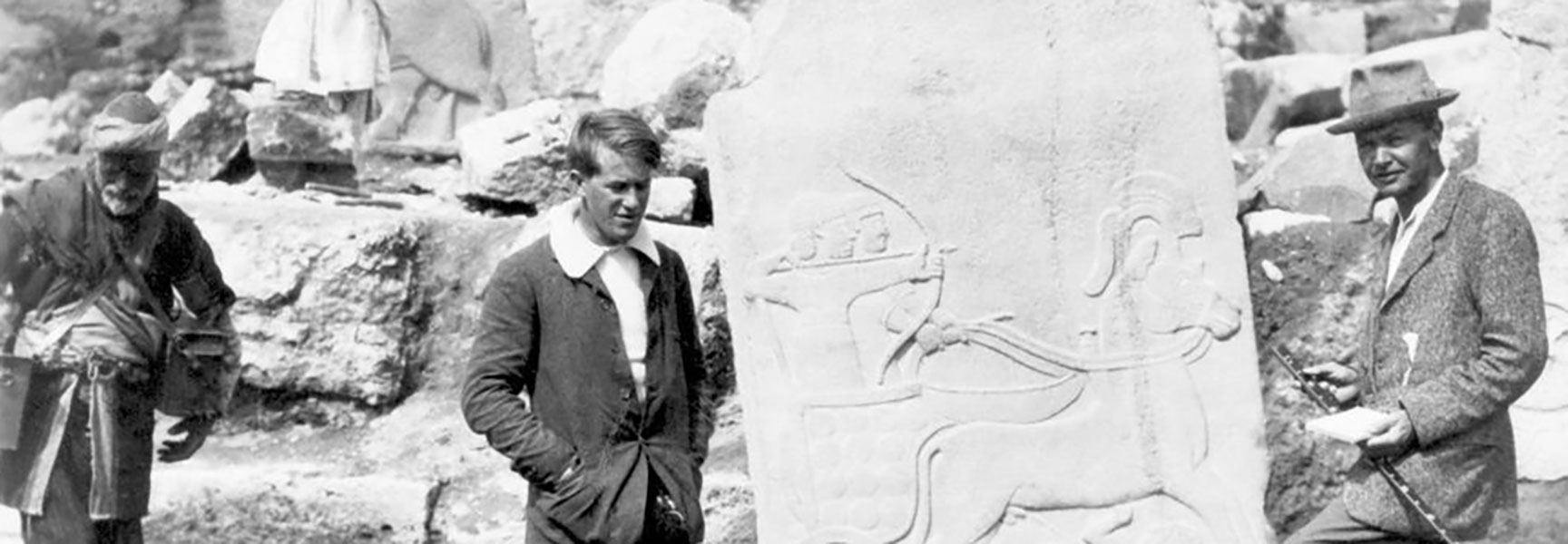 A black and white photograph shows T. E. Lawrence and Leonard Woolley examining a large, carved stone at an archaeological dig.