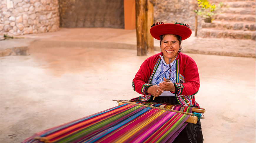 A smiling Peruvian woman in traditional clothing sits at a loom, weaving a colorful textile in a rustic courtyard.