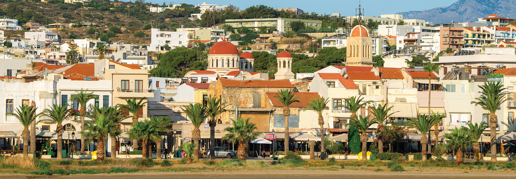 A scenic view of a coastal town in Greece, featuring a red-domed church, hillside buildings, and a waterfront lined with palm trees.