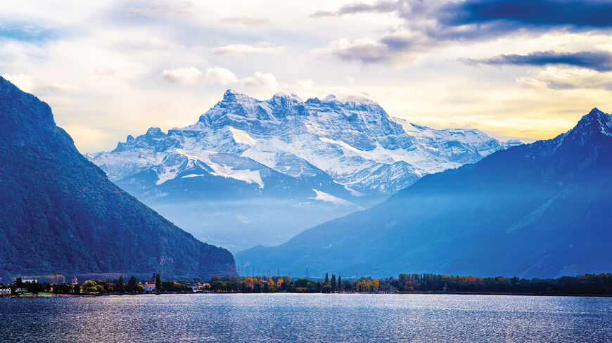 Scenic view of Lake Geneva with snow-capped mountains of the Swiss and French Alps in the background under a dramatic sky.