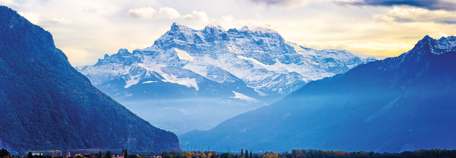 Scenic view of Lake Geneva with snow-capped mountains of the Swiss and French Alps in the background under a dramatic sky.