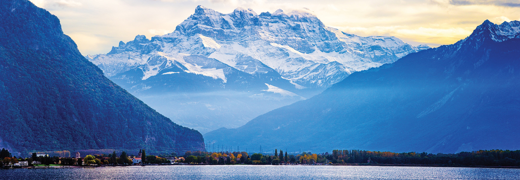 Snow-covered peaks of the Dents du Midi mountain range tower over the tree-lined shore of Lake Geneva in Switzerland and France.
