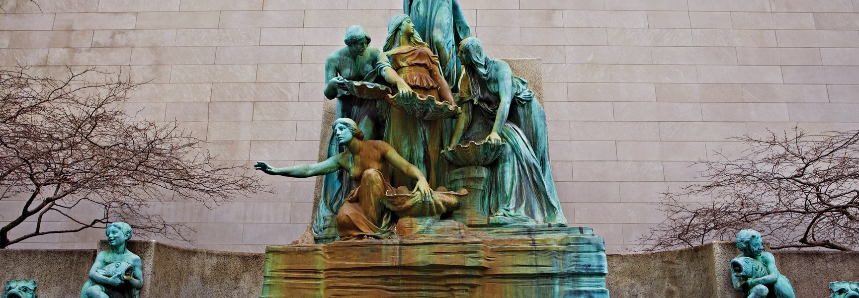 A large, weathered green statue of several figures holding shells stands before a stone wall in the Netherlands.