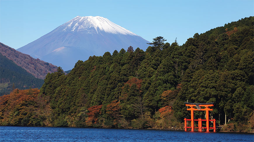 A red torii gate stands in Lake Ashi with snow-capped Mount Fuji visible in the background behind a dense forest.