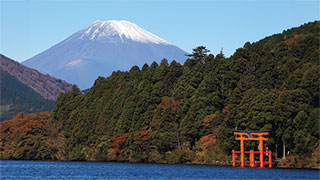 A snow-capped Mount Fuji rises behind a forested shoreline, with a red torii gate standing in the calm blue water of Lake Ashi.