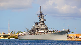 The battleship USS Missouri is docked in a harbor under a partly cloudy sky.