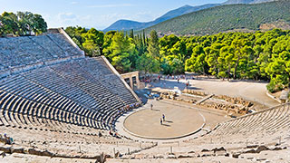 The ancient stone Theater of Epidaurus is nestled into a green hillside, with a few tourists on the circular stage below.