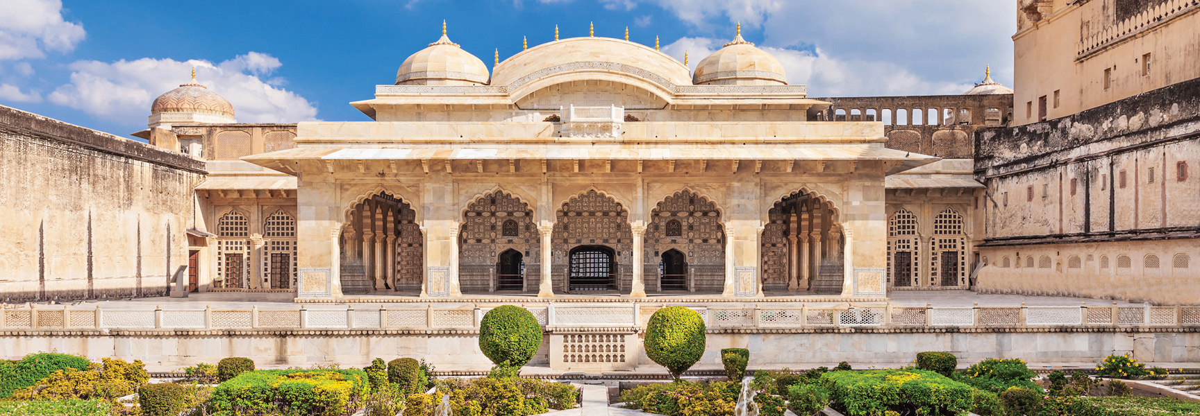 An ornate, tan-colored palace with domes and arches sits behind a manicured garden under a blue sky in India.