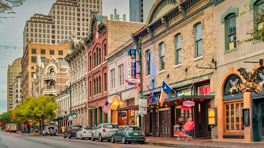Historic buildings with neon signs line the vibrant Sixth Street entertainment district in Austin, Texas, with cars parked along the busy road.