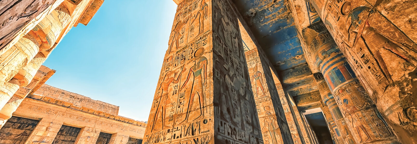 Intricate hieroglyphics and ancient carvings adorn massive stone pillars within an Egyptian temple ruin under a clear blue sky.