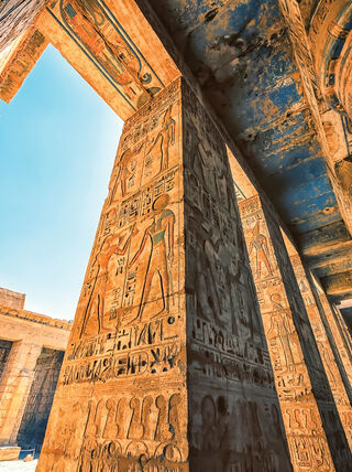 Intricate hieroglyphics and ancient carvings adorn massive stone pillars within an Egyptian temple ruin under a clear blue sky.