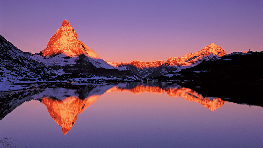 The snow-capped peak of the Matterhorn mountain in Switzerland reflects onto a still lake at sunrise.