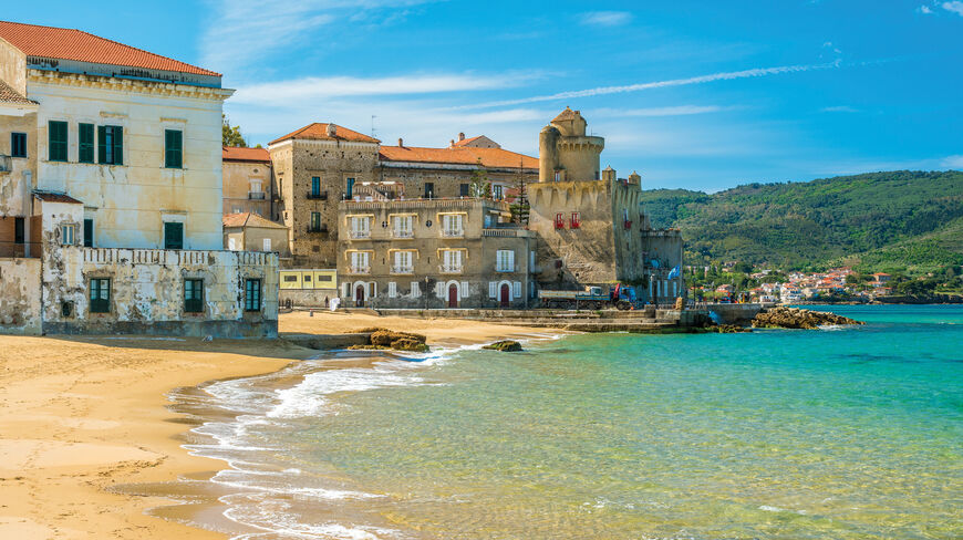 A peaceful beach with turquoise water in Cilento, Italy, lined with historic stone buildings and green hills under a bright blue sky.