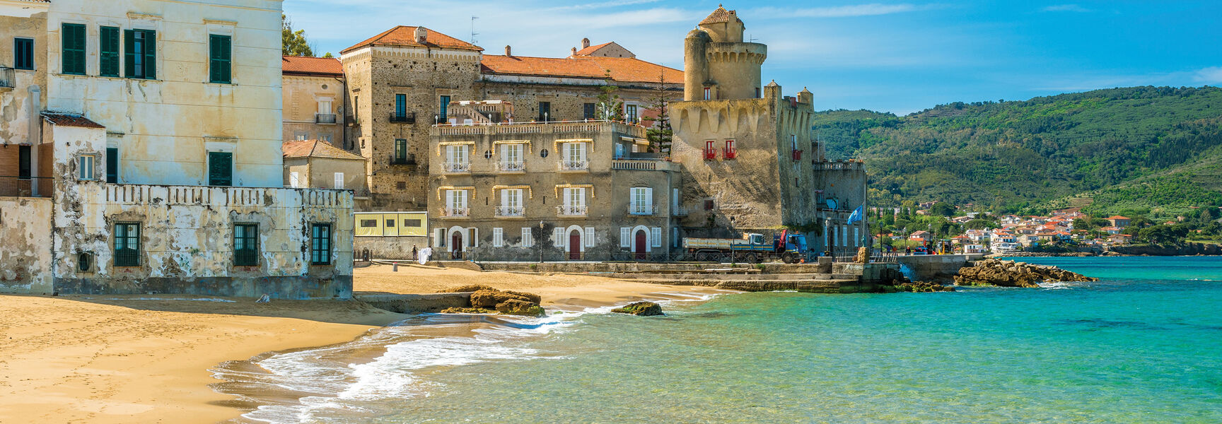 A peaceful beach with turquoise water in Cilento, Italy, lined with historic stone buildings and green hills under a bright blue sky.