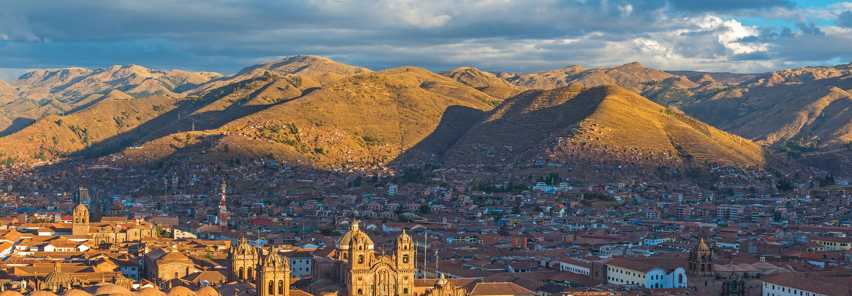 High-angle landscape of Cusco, Peru, featuring red-tiled roofs and historic cathedrals nestled in a valley surrounded by sunlit, rolling mountains under a cloudy sky.
