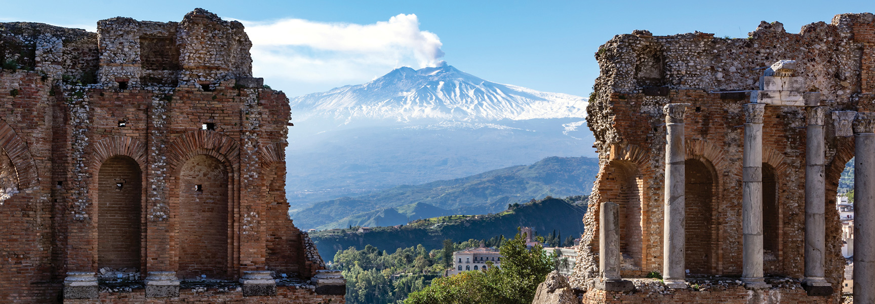 The ancient ruins of a theater in Sicily frame a view of the snow-capped, smoking Mount Etna in the distance under a blue sky.