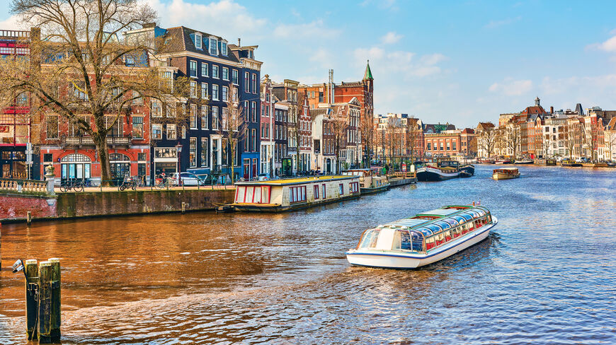 A tour boat travels down an Amsterdam canal lined with historic gabled buildings and houseboats under a clear blue sky.