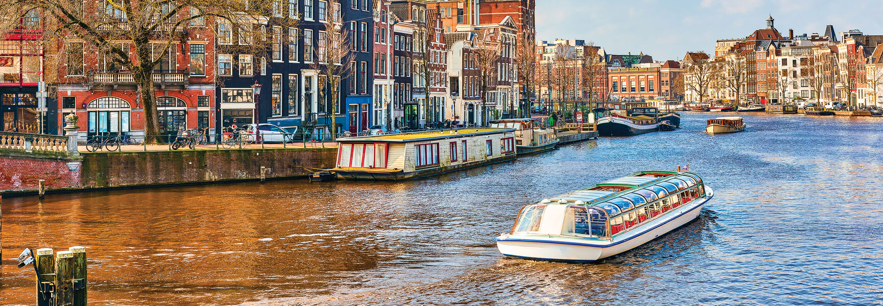 A tour boat travels down an Amsterdam canal lined with historic gabled buildings and houseboats under a clear blue sky.