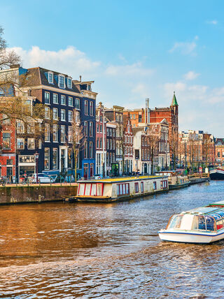 A tour boat travels down an Amsterdam canal lined with historic gabled buildings and houseboats under a clear blue sky.