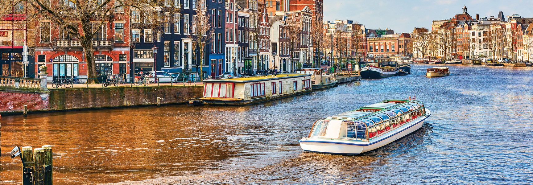 A tour boat travels down a canal in Amsterdam, past traditional canal houses and houseboats on a sunny day.